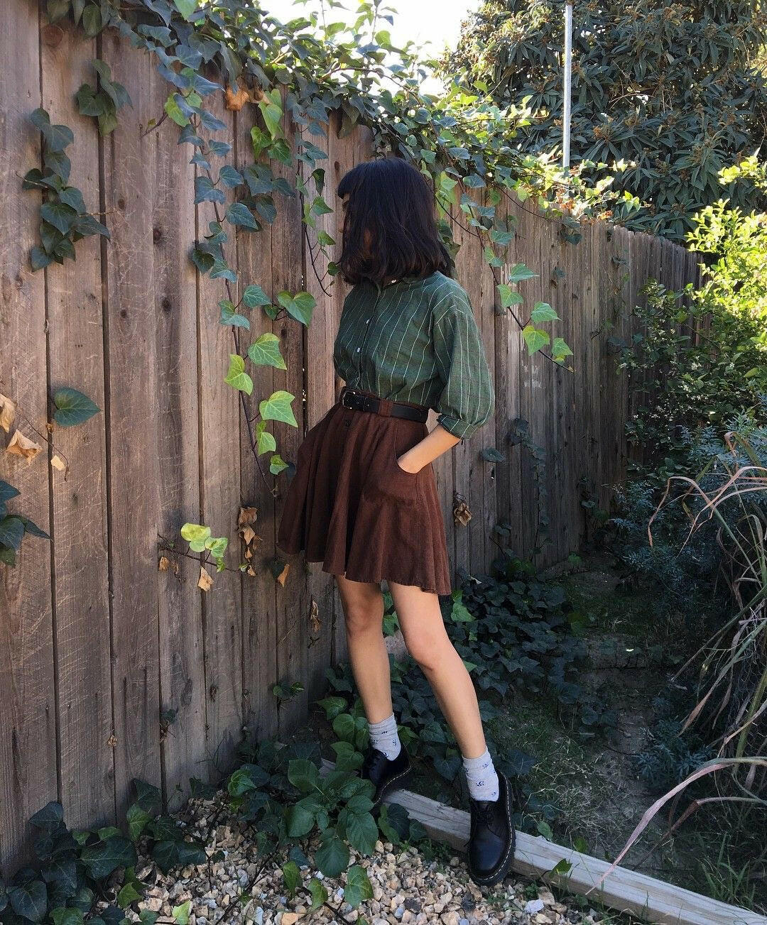 Person wearing a green shirt and brown skirt standing against a wooden fence with greenery.