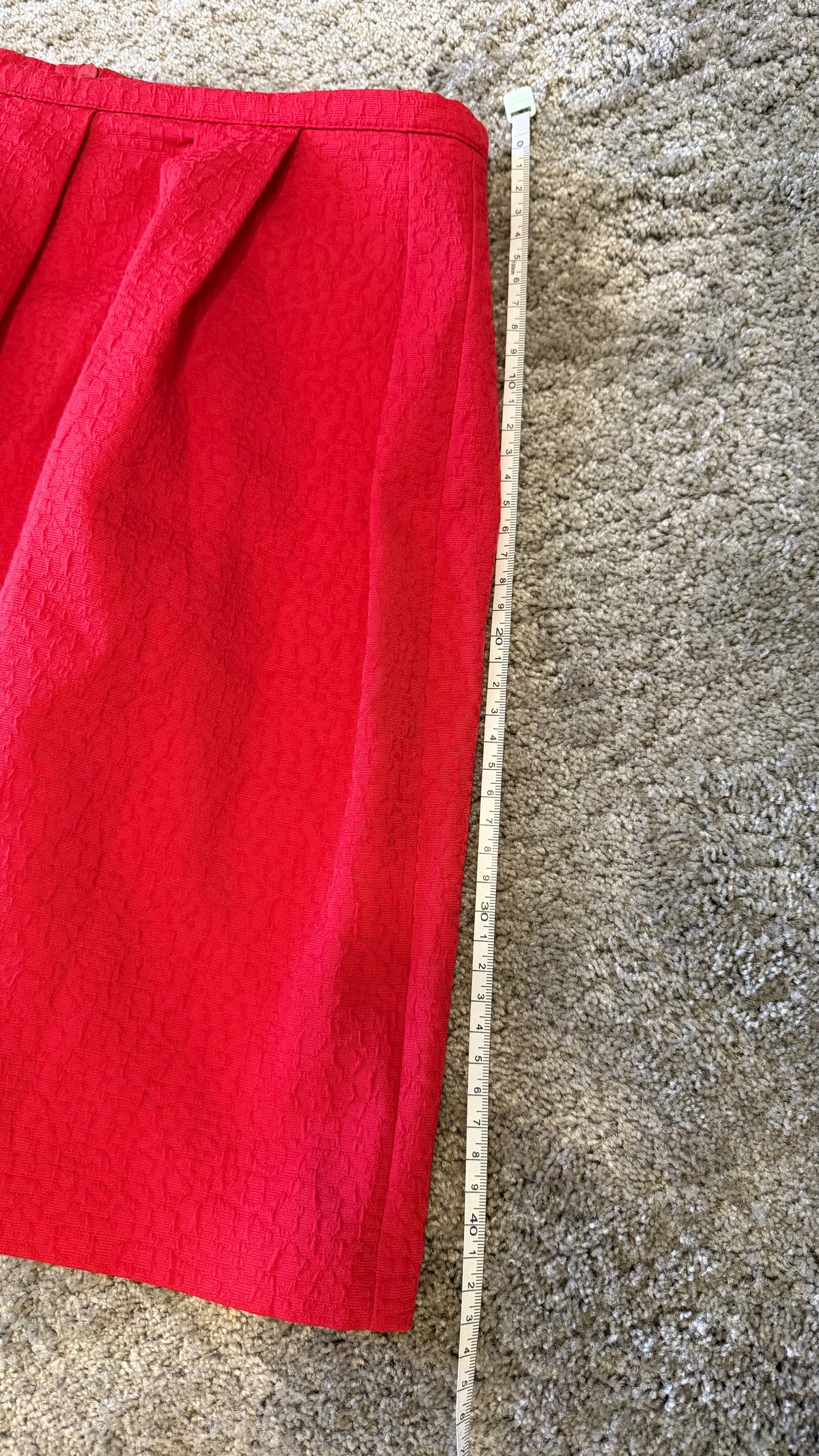 A bright red skirt laid flat on a beige carpet next to a wooden tape measure to show the length.