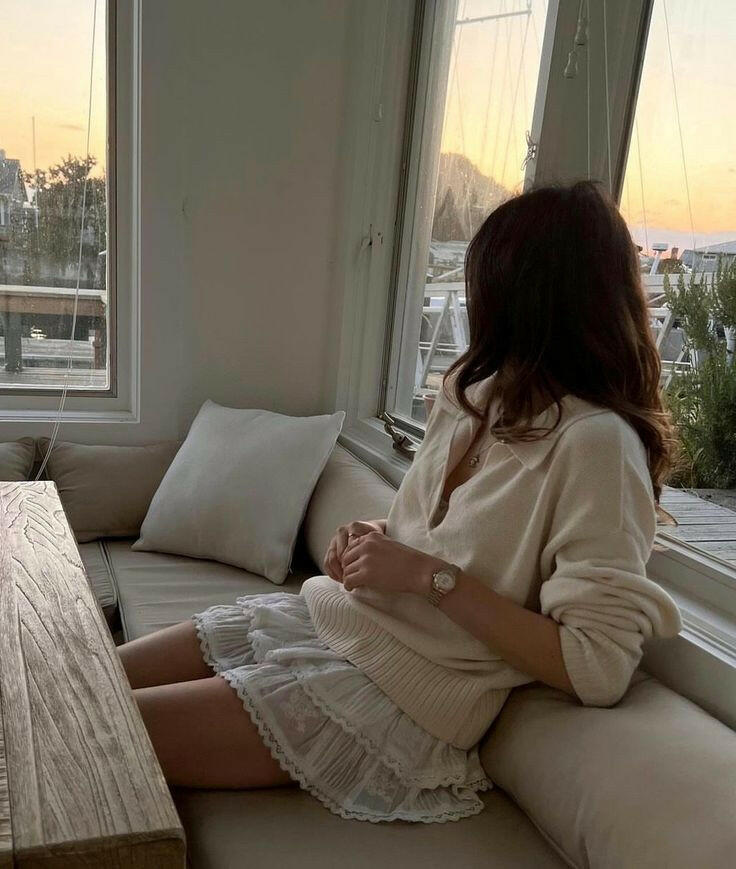 Woman sitting on a couch by a window, looking out at a cityscape during sunset.
