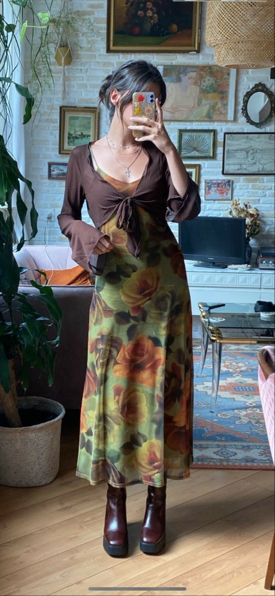 A woman takes a mirror selfie in a cozy, decorated room, wearing a brown long-sleeve top layered over a long-sleeved maxi dress with a green, yellow, and orange floral print, paired with dark brown boots.