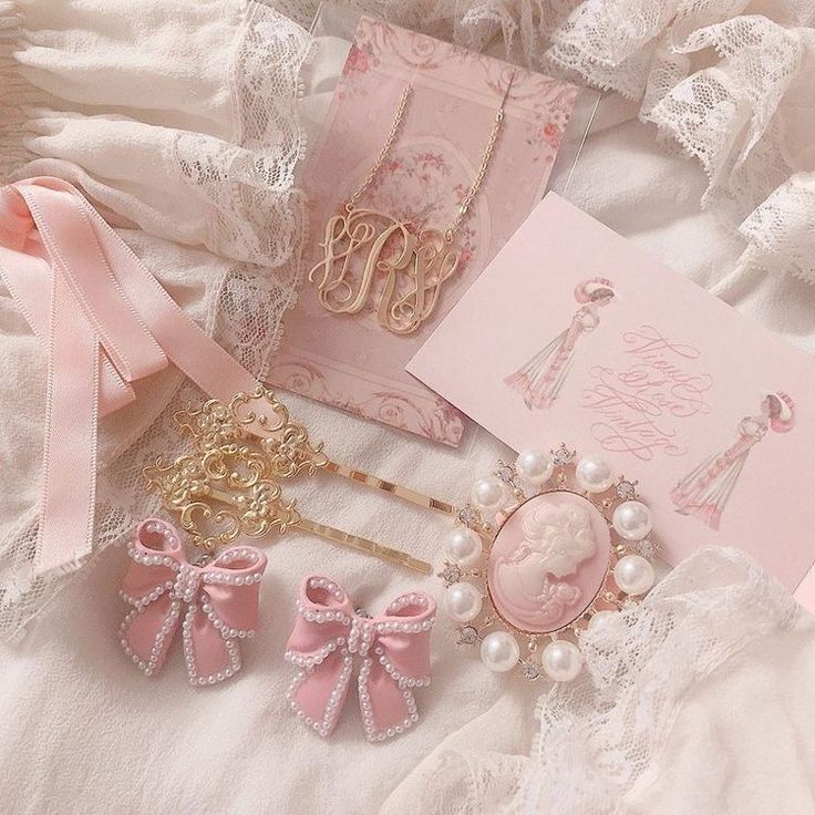 A styled flat-lay of pink and gold jewelry accessories on a soft white lace fabric background, including a monogram necklace on a card, a brooch with a cameo-style pink face and pearl border, two pink bow hair clips with pearl trim, and a decorative hairpin.