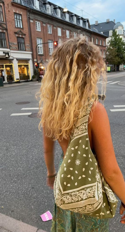 A woman with long, curly blonde hair seen from behind, wearing a green and beige patterned bandana shoulder bag and a matching green patterned dress while standing on a city street.