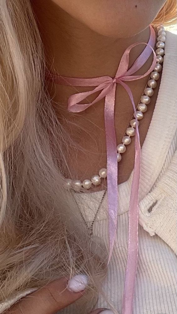 A close-up of a person wearing a pink satin ribbon choker with a bow and dangling ribbon ends, layered with a pearl necklace and a delicate silver chain.