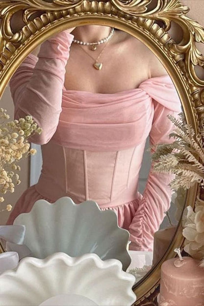 A person wearing a light pink off-the-shoulder dress with long sleeves and a structured corset-style bodice, photographed in front of an ornate gold mirror. The scene includes decorative dried flowers, a white scalloped dish, and a pink candle.