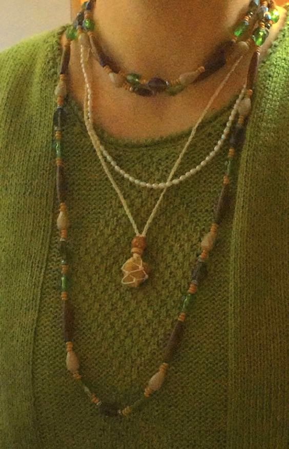 A close-up of a person wearing a layered necklace set over an olive green knit sweater. The set includes a short beaded choker, a long beaded necklace with a small shell pendant, and a thin white beaded strand.