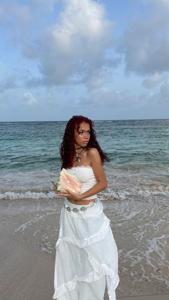 A woman with curly red hair stands on a sandy beach holding a large conch shell, wearing a white strapless top and a long white skirt with silver shell-like embellishments. The ocean and cloudy sky are in the background.