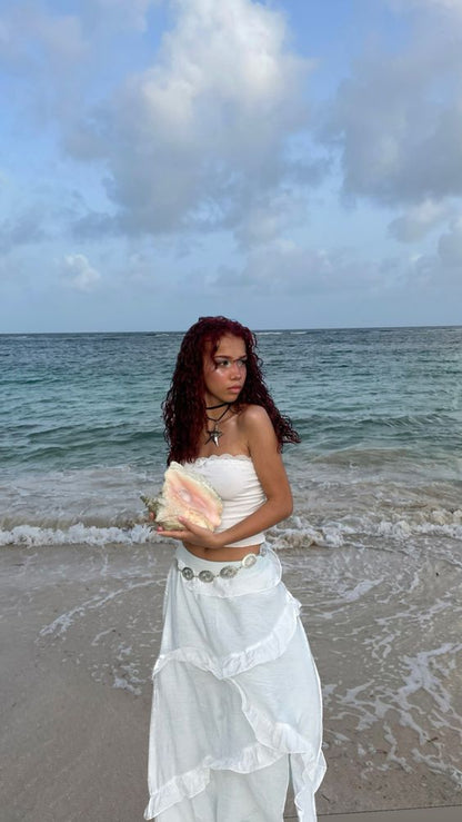 A woman with curly red hair stands on a sandy beach holding a large conch shell, wearing a white strapless top and a long white skirt with silver shell-like embellishments. The ocean and cloudy sky are in the background.