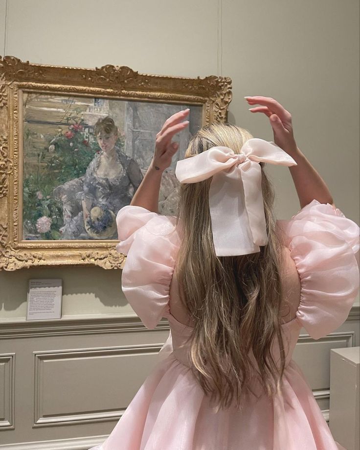 A woman with long blonde hair wearing a light pink dress and a large matching bow in her hair, photographed from behind as she looks at a framed painting in a gallery.