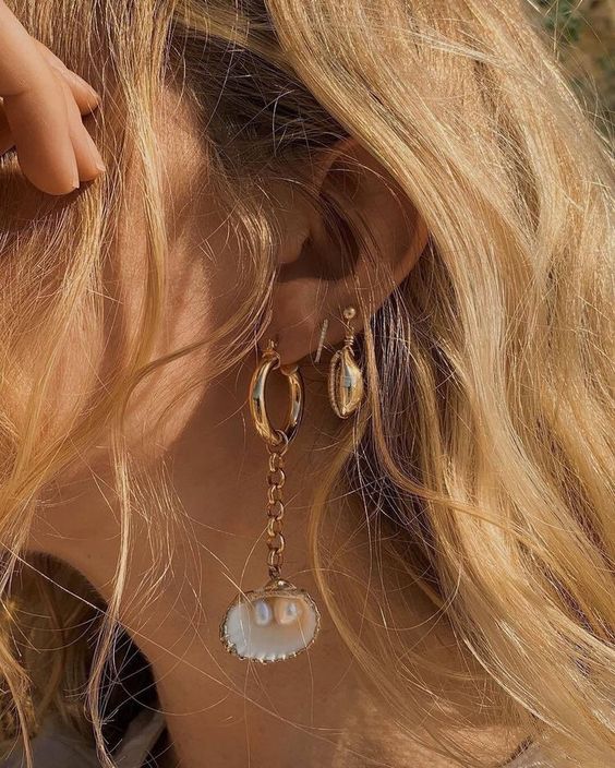 A close-up of a woman's ear wearing a gold hoop earring with a dangling chain and a white shell-shaped charm, styled with wavy blonde hair in warm natural light.