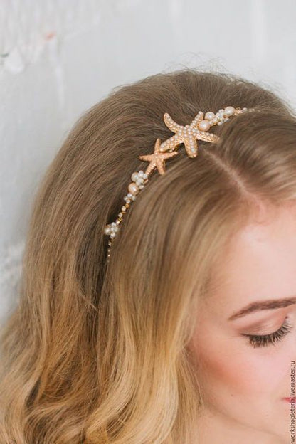 A close-up of a woman with blonde hair wearing a delicate gold headband decorated with two starfish and small pearl-like beads, set against a light textured wall.