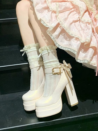 A pair of chunky platform high-heeled boots in glossy off-white, styled with cream ribbed lace socks and a light pink ruffled skirt on a model sitting on black steps.