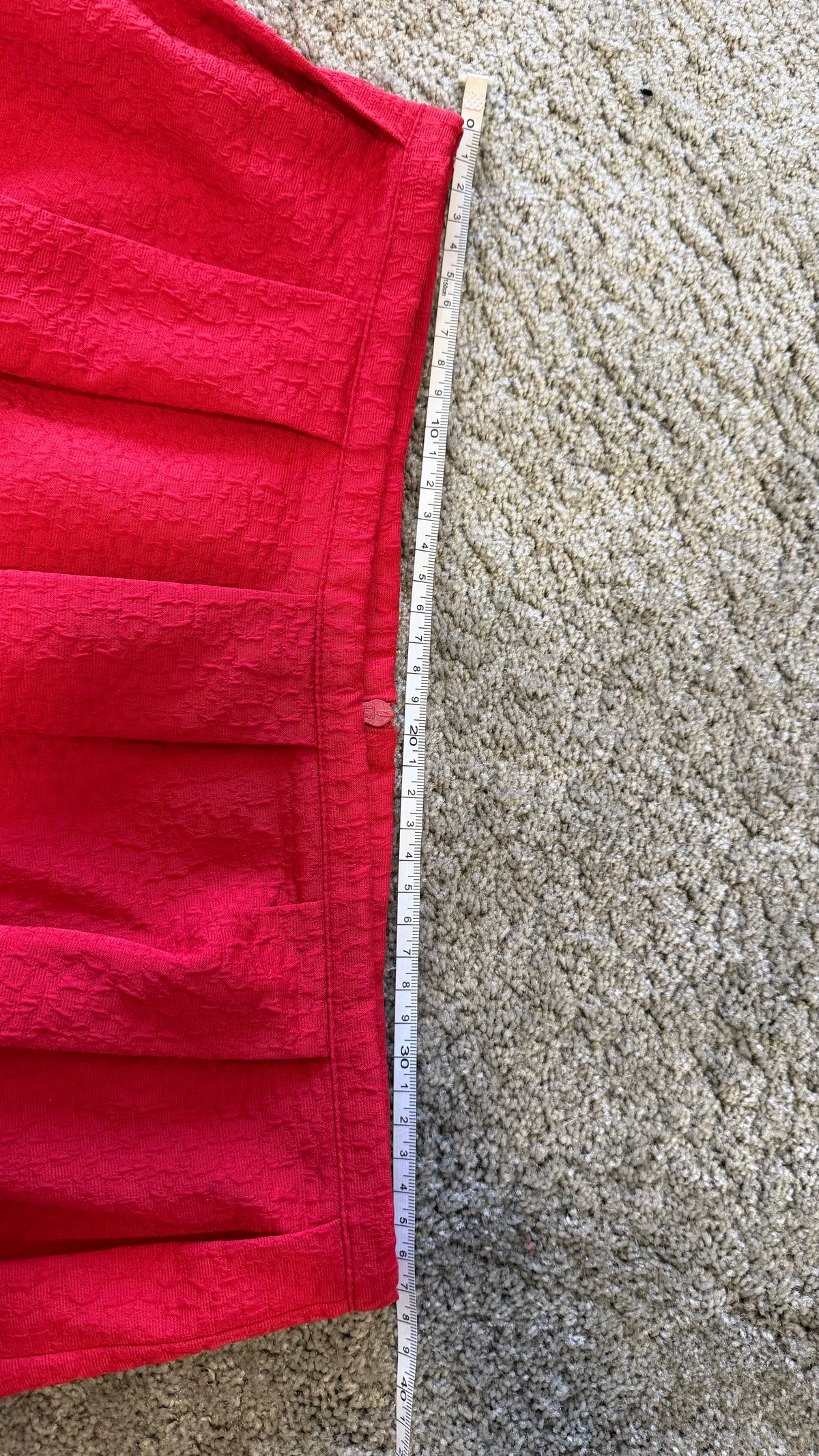 A bright red pleated fabric garment laid flat on a beige carpet next to a measuring tape to show dimensions.