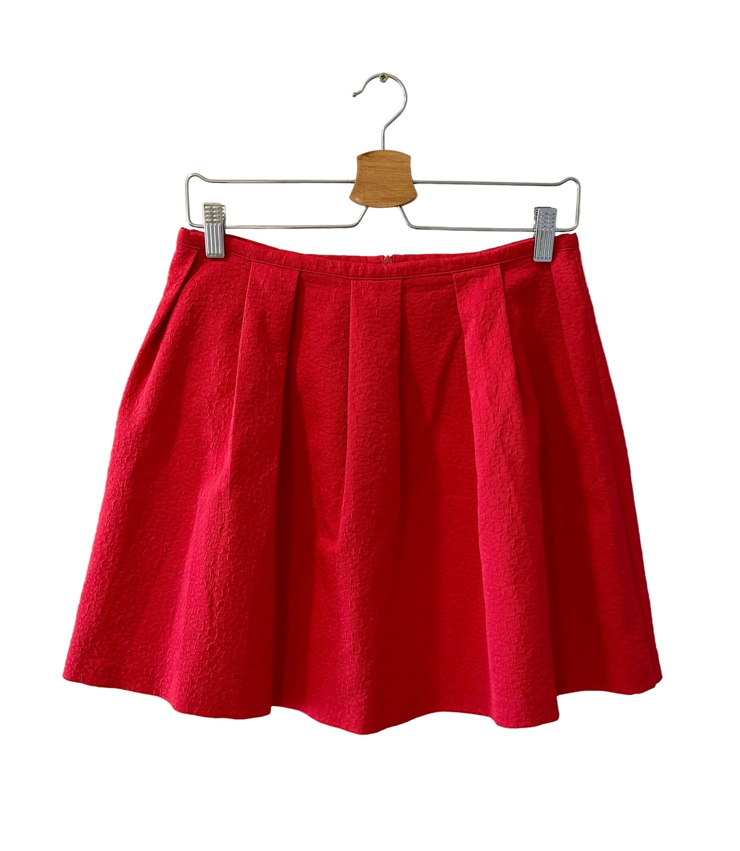 A bright red pleated mini skirt hanging from a metal hanger with a wooden brand tag, clipped to a wire hanger against a plain off-white wall.