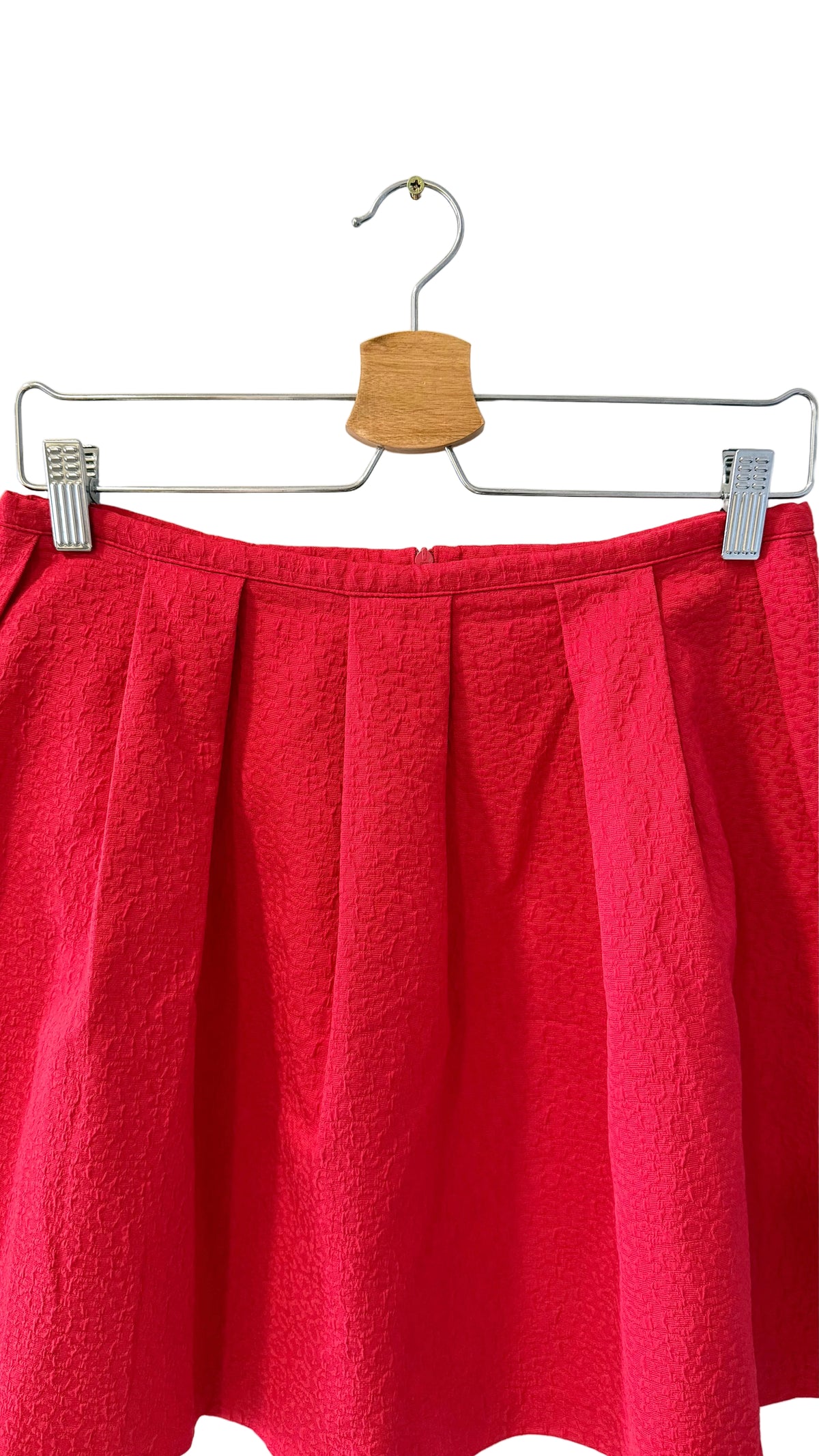 A bright red pleated skirt hanging from a metal hanger with a wooden cone-shaped hook against a plain off-white wall.