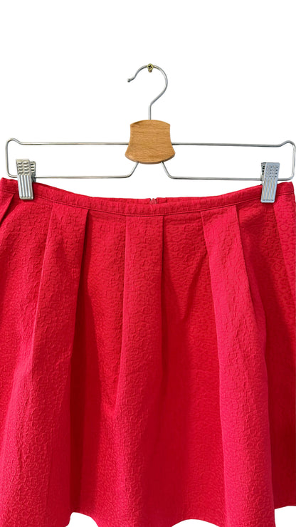 A bright red pleated skirt hanging from a metal hanger with a wooden cone-shaped hook against a plain off-white wall.
