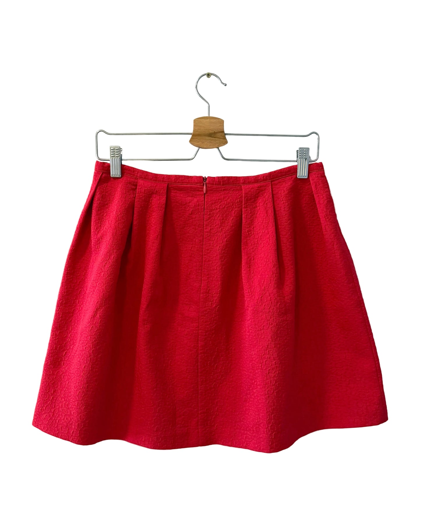 A bright red pleated mini skirt hanging from a metal hanger with a wooden clip against a plain white wall.