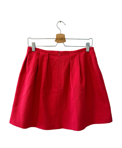 A bright red pleated mini skirt hanging from a metal hanger with a wooden clip against a plain white wall.