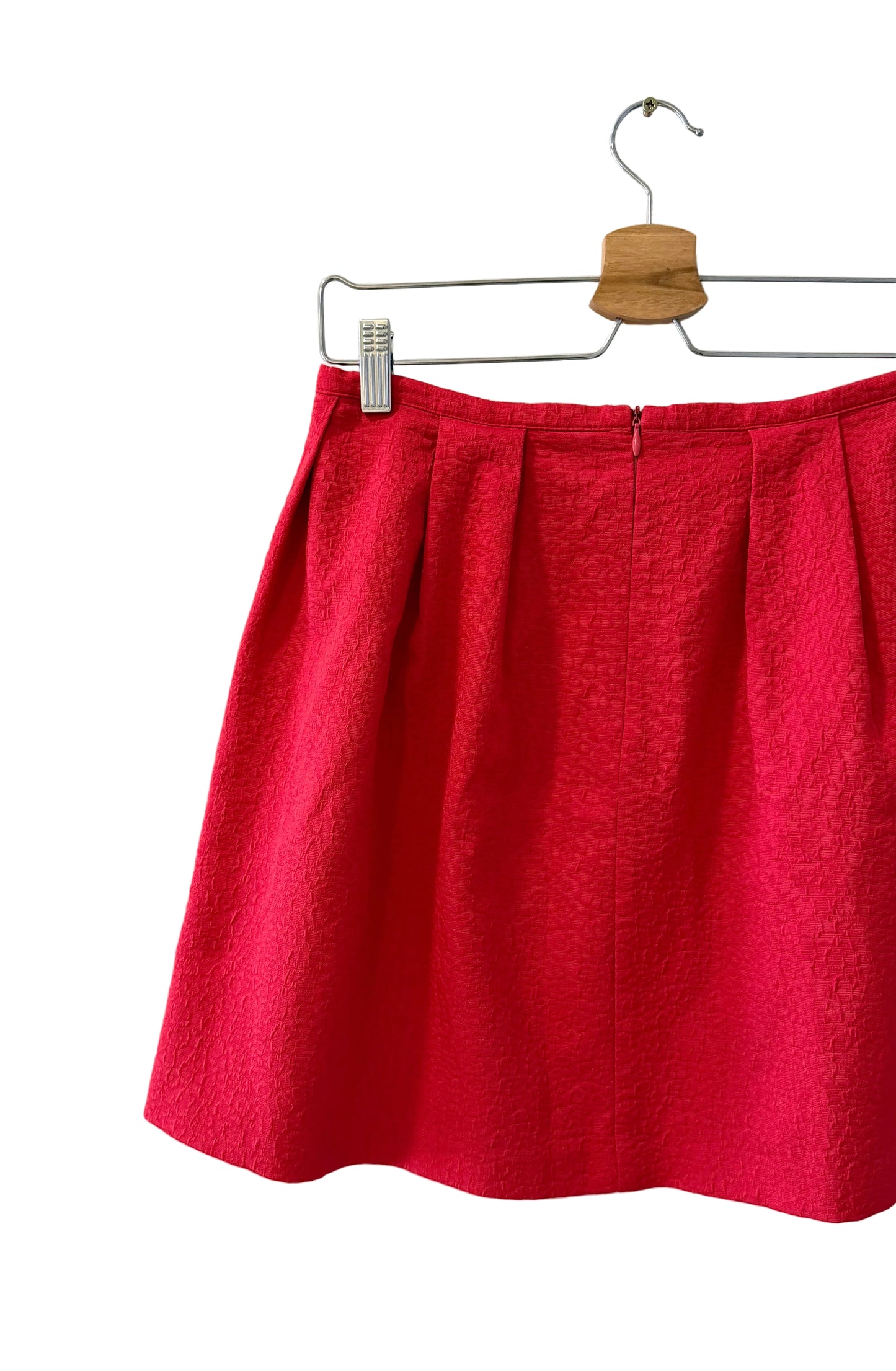 A bright red textured pleated skirt hanging from a metal hanger against a plain off-white wall.