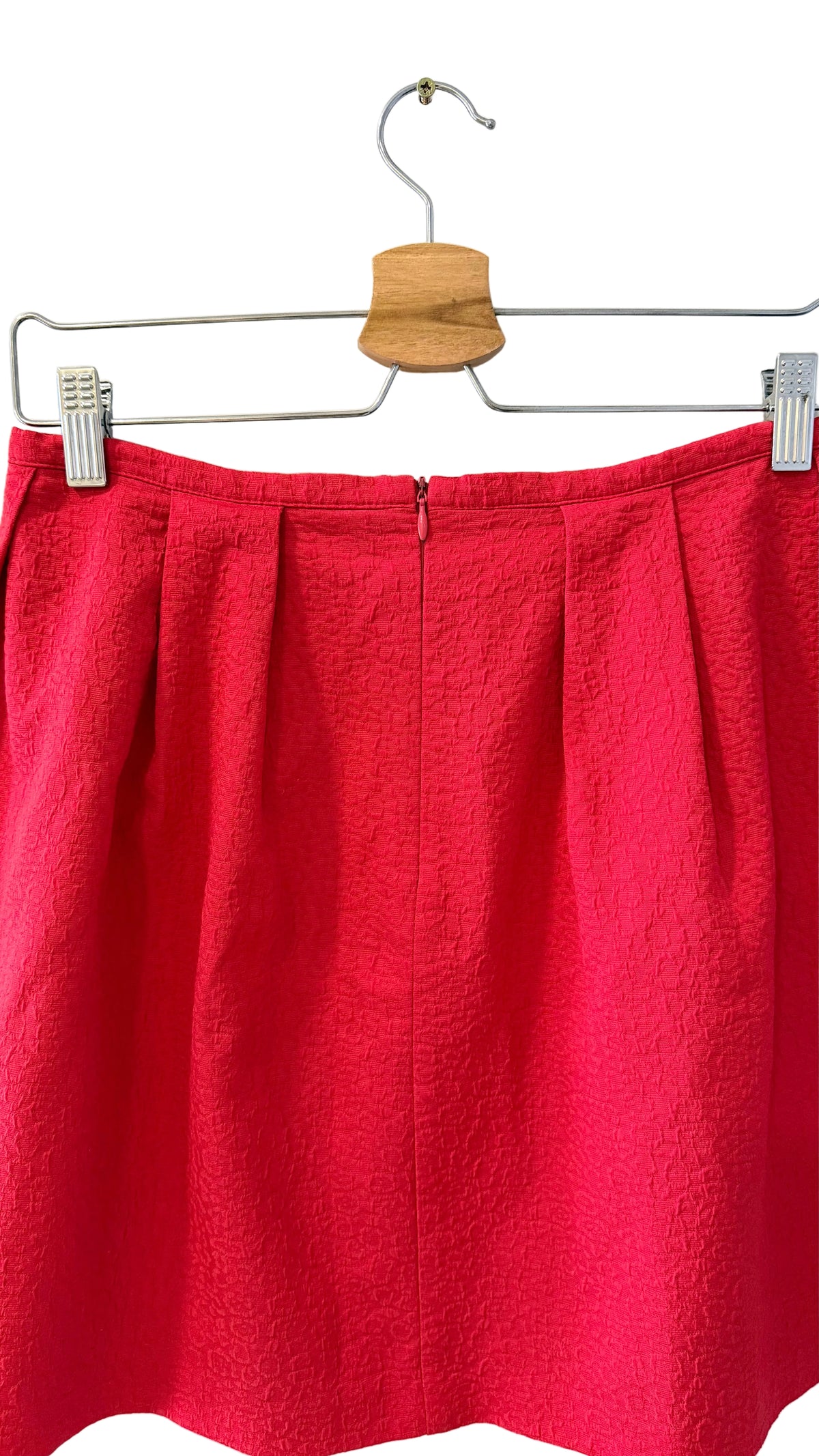 A bright red pleated skirt hanging from a wooden hanger with metal clips against a plain white wall.