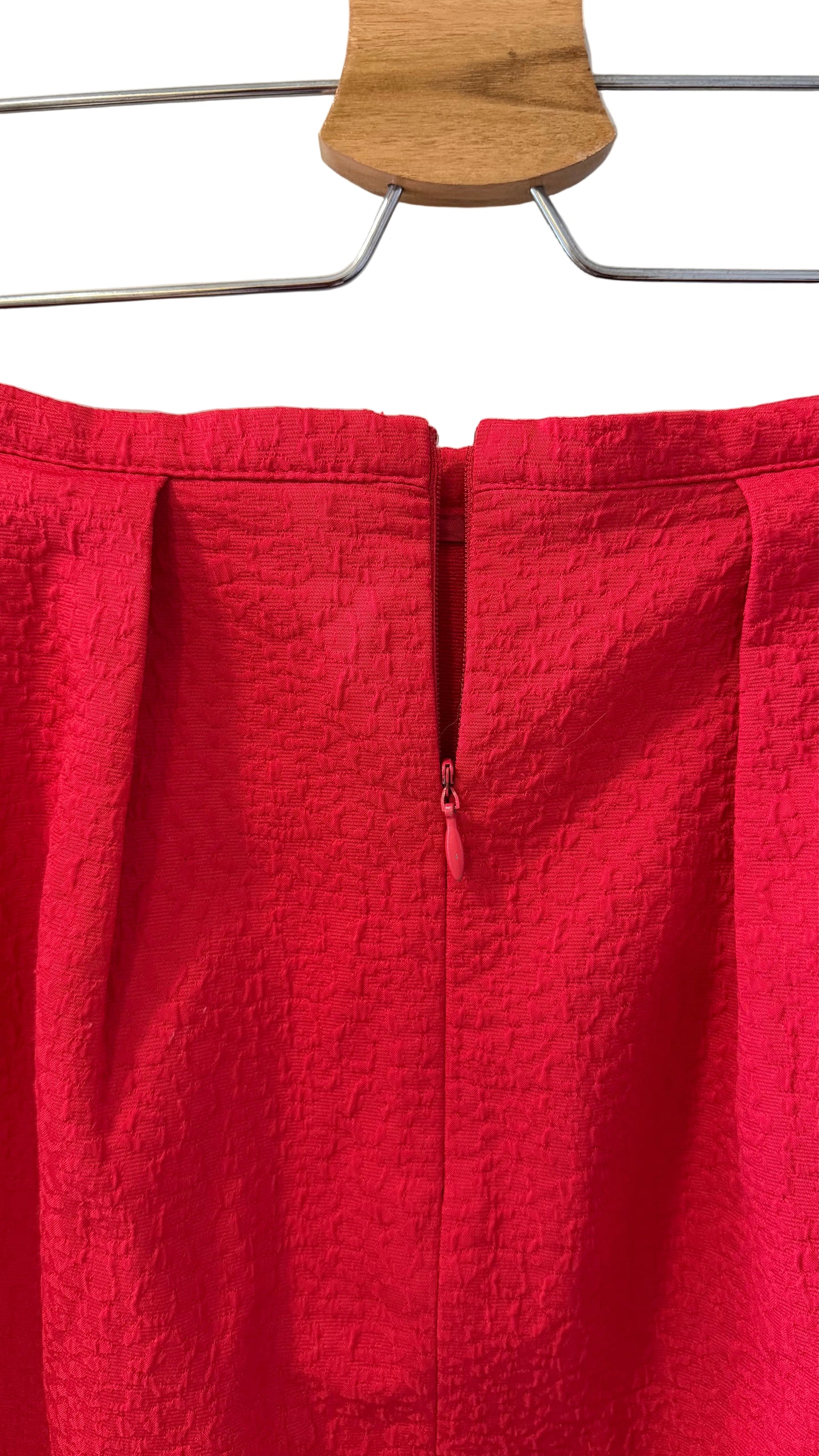 A close-up of a bright red textured skirt hanging on a wooden hanger against a white wall.