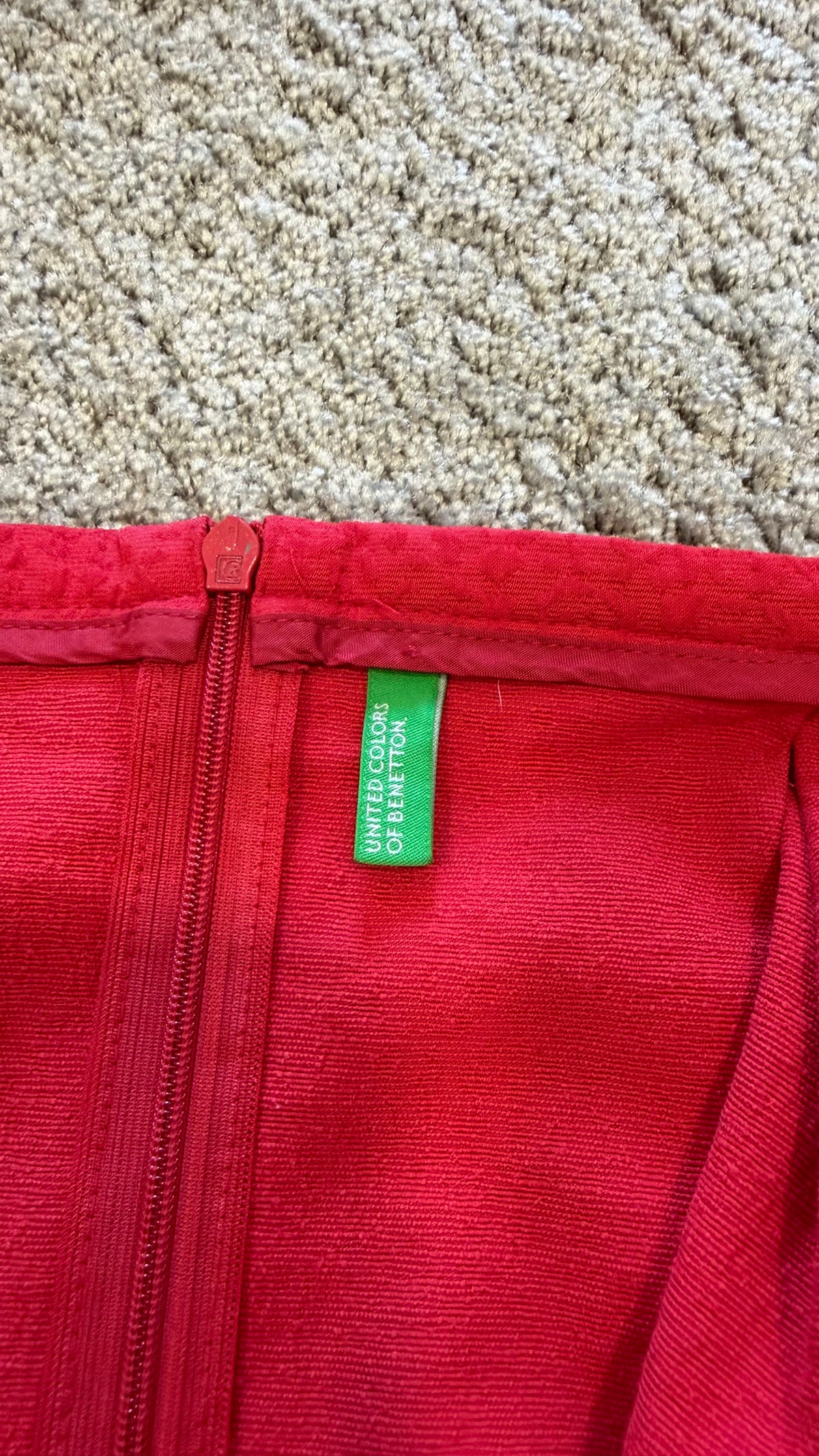 Close-up of a bright red zippered garment laid flat on a beige carpet, showing a small green interior brand tag that reads 'DROPSHOPS OF BIRMINGHAM'.