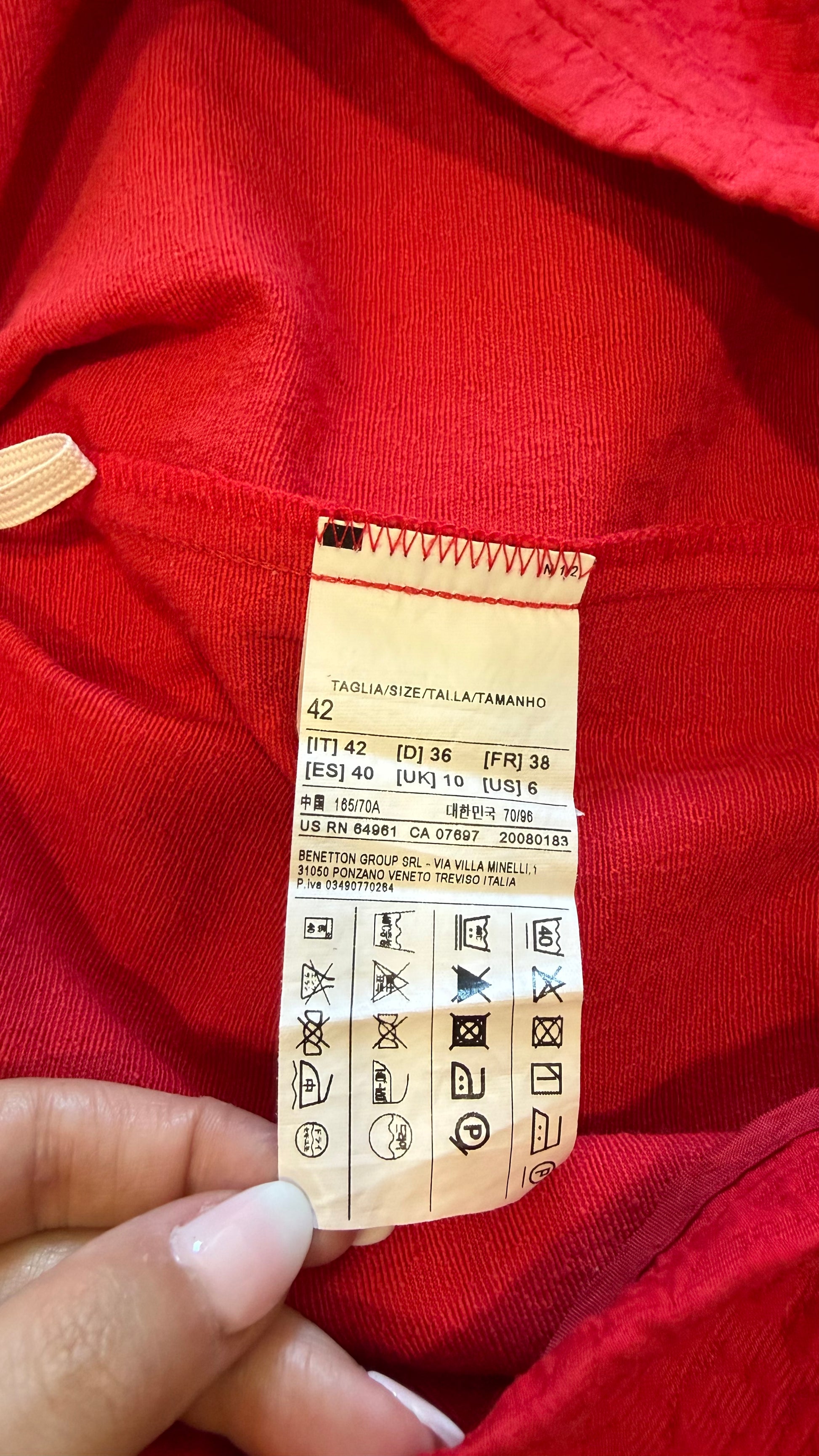 A close-up of a hand holding a fabric care label attached to a bright red knit garment.
