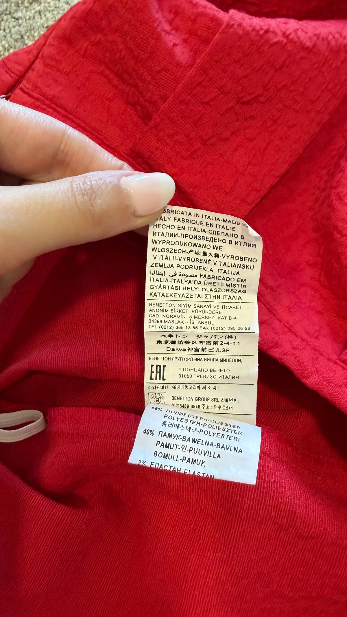 A close-up of a red garment with a hand holding a sewn-on care label and a separate white EHC authenticity tag attached below it.