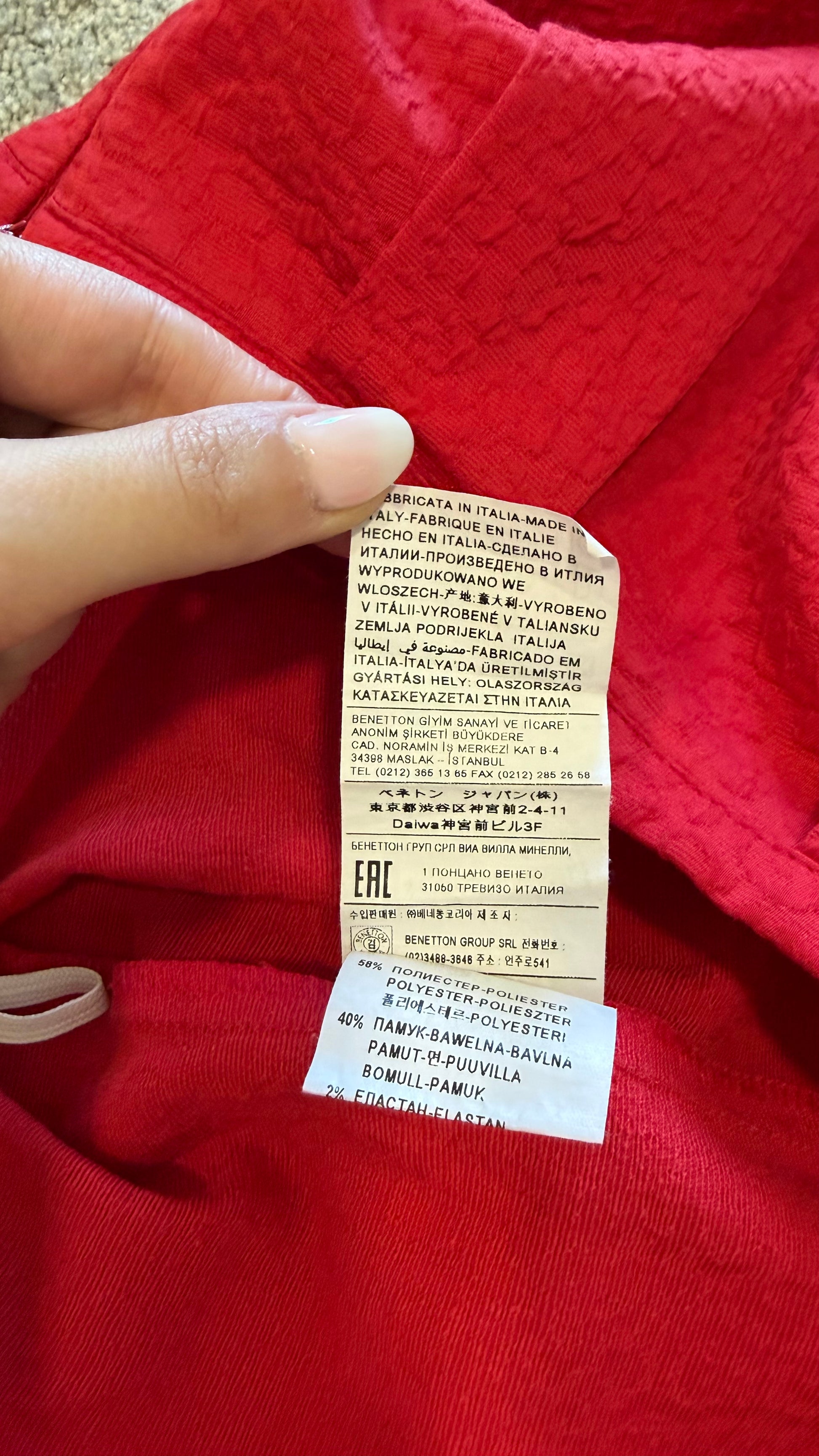 A close-up of a red garment with a hand holding a sewn-on care label and a separate white EHC authenticity tag attached below it.