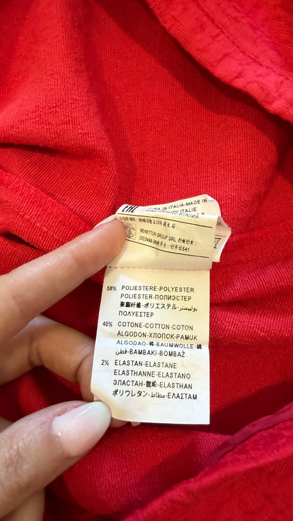A close-up of a hand holding a fabric care label attached to a bright red knit garment.