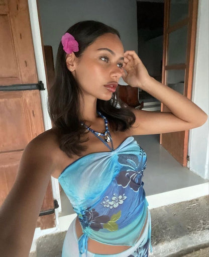A woman wearing a blue floral halter top and matching bottoms, posing in front of a doorway with a pink flower in her hair.