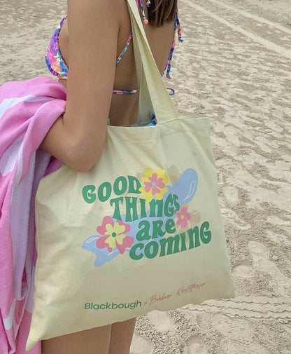 A cream canvas tote bag with colorful retro-style text reading 'GOOD THINGS ARE COMING' and floral graphics, carried on a beach by a person wearing a multicolor bikini and holding a pink towel. The bag has 'Blackbough' printed near the bottom.
