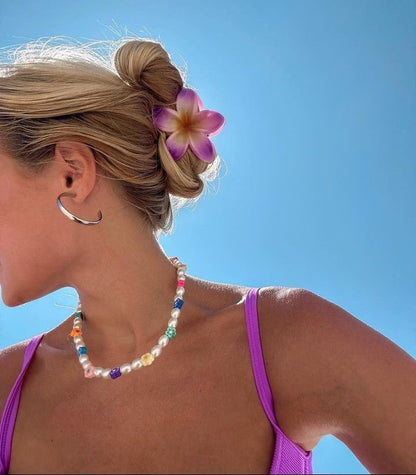 A close-up side profile of a woman outdoors against a clear blue sky, wearing a colorful beaded necklace with white pearls and bright multicolor beads, along with a purple flower hair clip and hoop earring.