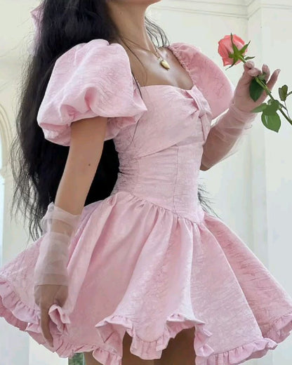 A woman wearing a light pink textured puff-sleeve dress with a sweetheart neckline, corset-style bodice, and flared ruffled skirt, holding a pink rose.