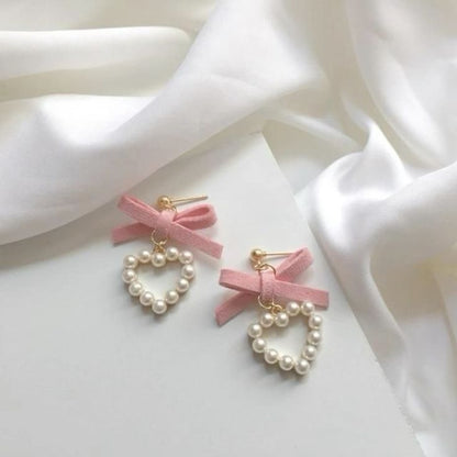 A pair of dangling earrings with pink ribbon bows, white faux pearls, and gold-tone posts, styled on a white surface with draped white fabric.