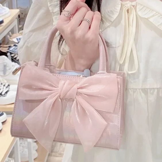 A person in a white ruffled top holds a shiny light pink handbag with a large bow on the front. The background shows a retail store interior with shoes and furniture.
