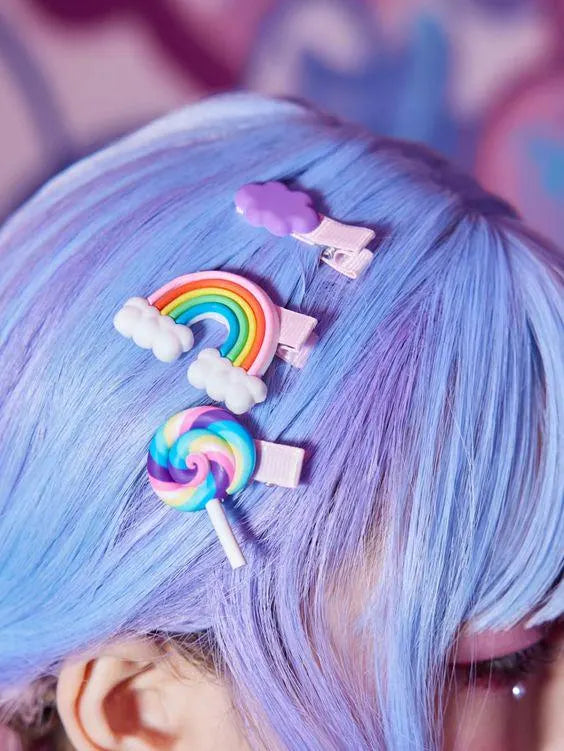 A close-up of pastel blue hair styled with three colorful hair clips: a rainbow clip with white clouds, a lollipop clip with rainbow swirls, and a small purple cloud clip.