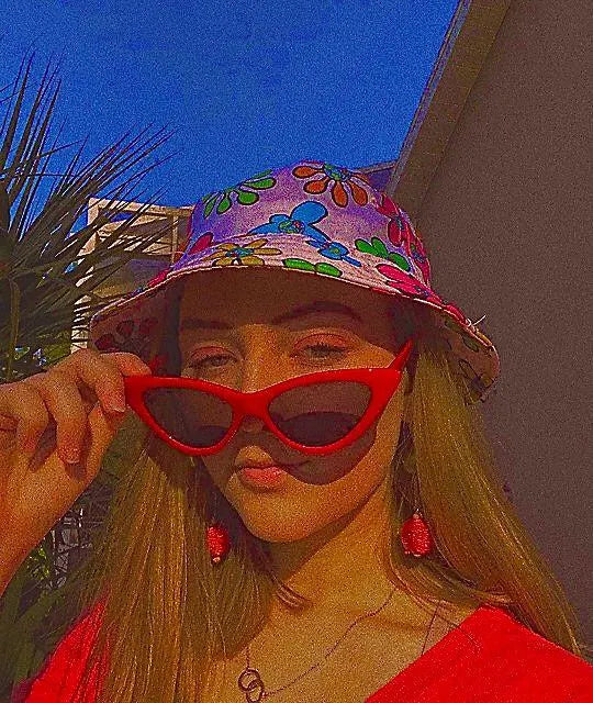 A woman outdoors wearing a colorful floral bucket hat, red cat-eye sunglasses, and red earrings, with a strong warm color cast over the photo.
