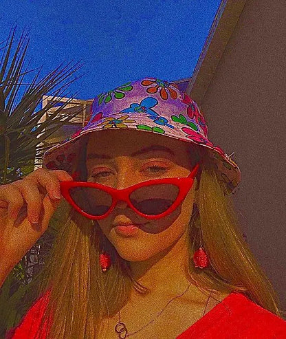 A woman outdoors wearing a colorful floral bucket hat, red cat-eye sunglasses, and red earrings, with a strong warm color cast over the photo.