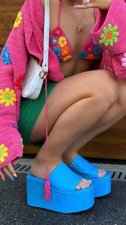 A person seated outdoors wearing a bright pink crocheted cardigan with colorful flower patches, a green bikini top, and vivid blue platform mule sandals. A white shoulder bag is also visible.
