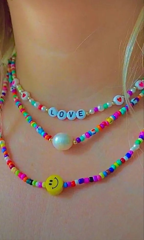 A close-up of a person wearing a multi-layered beaded necklace set. The necklaces feature colorful round beads, white pearl-like beads, a yellow smiley face charm, and a light blue letter charm spelling 'LOVE'.