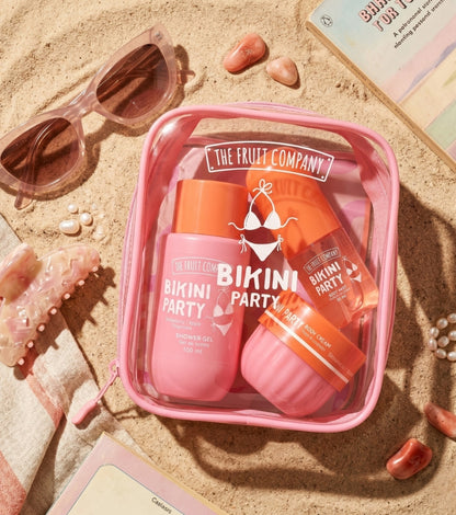 A pink zippered cosmetic bag filled with pink and orange swim-themed products from The Fruit Company, styled on sand with sunglasses, shells, and beach accessories.